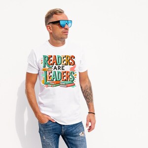 May include: White t-shirt with the text "READERS ARE LEADERS" in colorful lettering. The design includes stacked books in shades of green, orange, and red. The shirt is worn by a person wearing sunglasses and blue jeans.