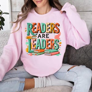 May include: A light pink sweatshirt with the text "READERS ARE LEADERS" in a colorful, layered design. The design includes stacked books in orange, green, and pink. The sweatshirt is a casual, comfortable style.