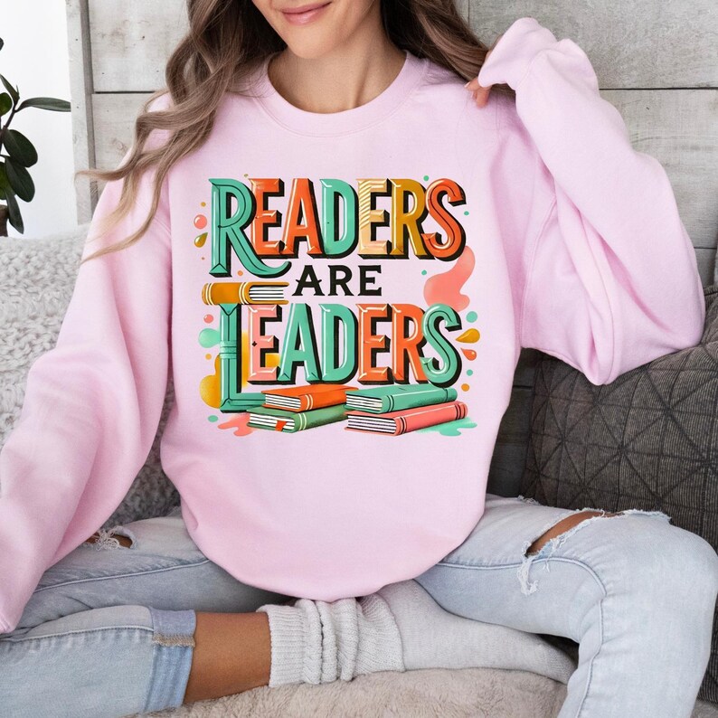 May include: A light pink sweatshirt with the text "READERS ARE LEADERS" in a colorful, layered design. The design includes stacked books in orange, green, and pink. The sweatshirt is a casual, comfortable style.