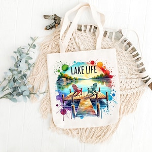 May include: A white canvas tote bag with a colorful watercolor design of a lake scene. The design features two chairs on a wooden dock, a sunset, and the text "LAKE LIFE".