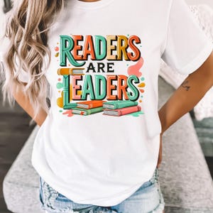 May include: White t-shirt with the text "READERS ARE LEADERS" in colorful, 3D lettering. The design includes stacks of books in green, orange, and pink. The shirt is a casual, everyday wear item.