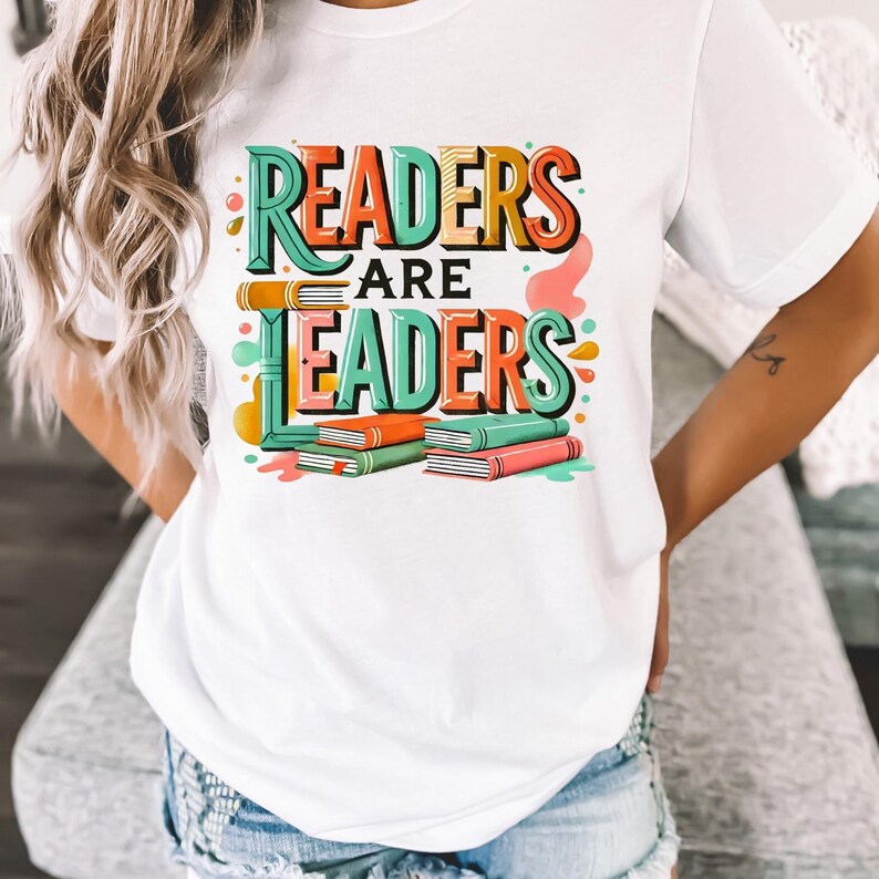 May include: White t-shirt with the text "READERS ARE LEADERS" in colorful, 3D lettering. The design includes stacks of books in green, orange, and pink. The shirt is a casual, everyday wear item.