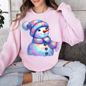 May include: A pink sweatshirt featuring a colourful cartoon snowman wearing a striped hat and scarf. The snowman has a sparkly, iridescent design.