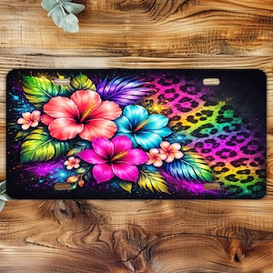 May include: Black license plate featuring vibrant hibiscus flowers in pink, red, and blue, with green and purple leaves. The background has a colorful leopard print design. The plate has two slots for mounting.