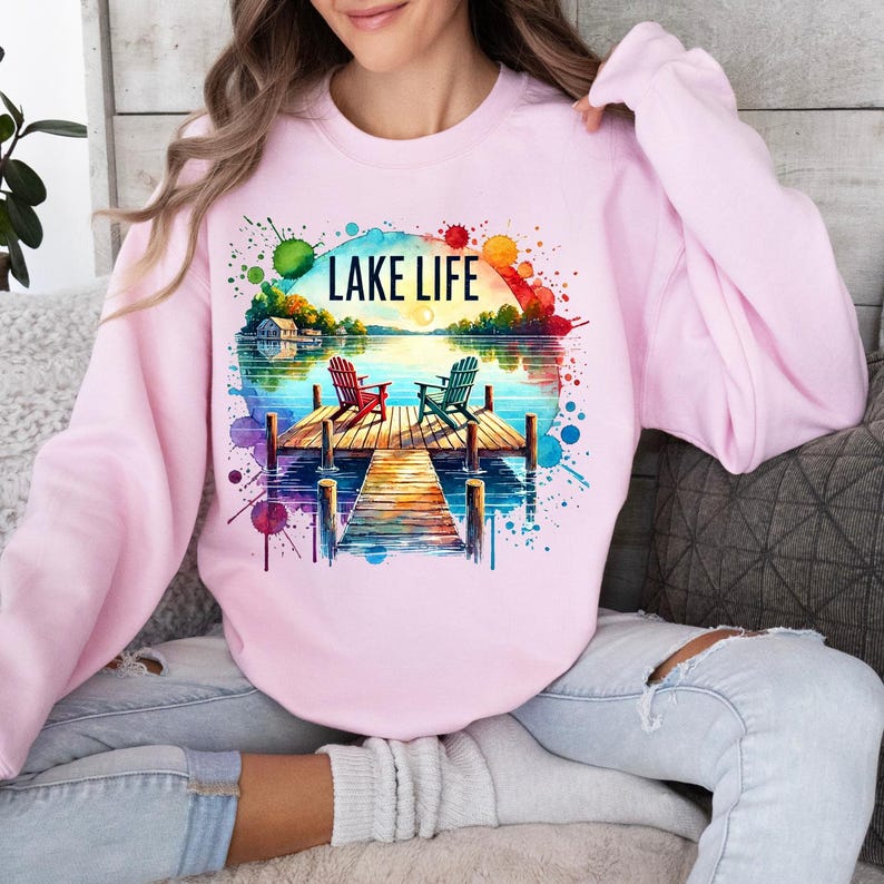 May include: Pink sweatshirt with a watercolor design of a lake scene with two chairs on a dock. The text "LAKE LIFE" is printed on the sweatshirt.