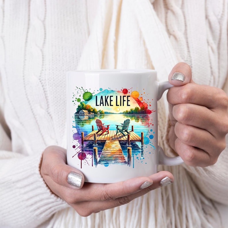 May include: White ceramic coffee mug with a watercolor design of a lake scene. The design features two chairs on a wooden dock, a lake with a sunset, and the text "LAKE LIFE".