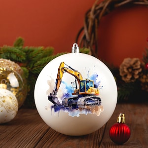 May include: White Christmas bauble with a watercolour illustration of a yellow excavator. The bauble has a silver cap and is displayed on a wooden surface with other festive decorations, including a red bauble and pine branches.