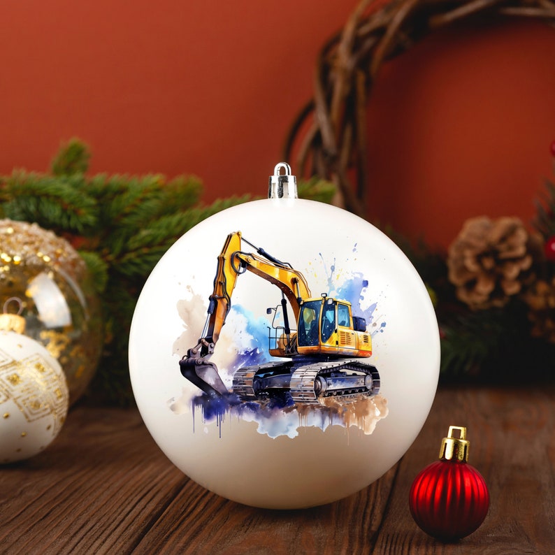 May include: White Christmas bauble with a watercolour illustration of a yellow excavator. The bauble has a silver cap and is displayed on a wooden surface with other festive decorations, including a red bauble and pine branches.