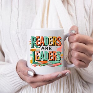 May include: White ceramic mug with the text "READERS ARE LEADERS" in colorful, stylized lettering. The design includes stacked books in orange, green, and pink. The mug is held by a person wearing a white sweater.