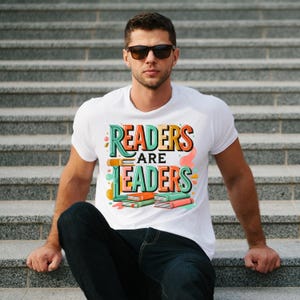 May include: White t-shirt with the text "READERS ARE LEADERS" in a colorful, retro design. The graphic includes stacked books in orange, green, and pink. The person is wearing sunglasses and sitting on gray steps.