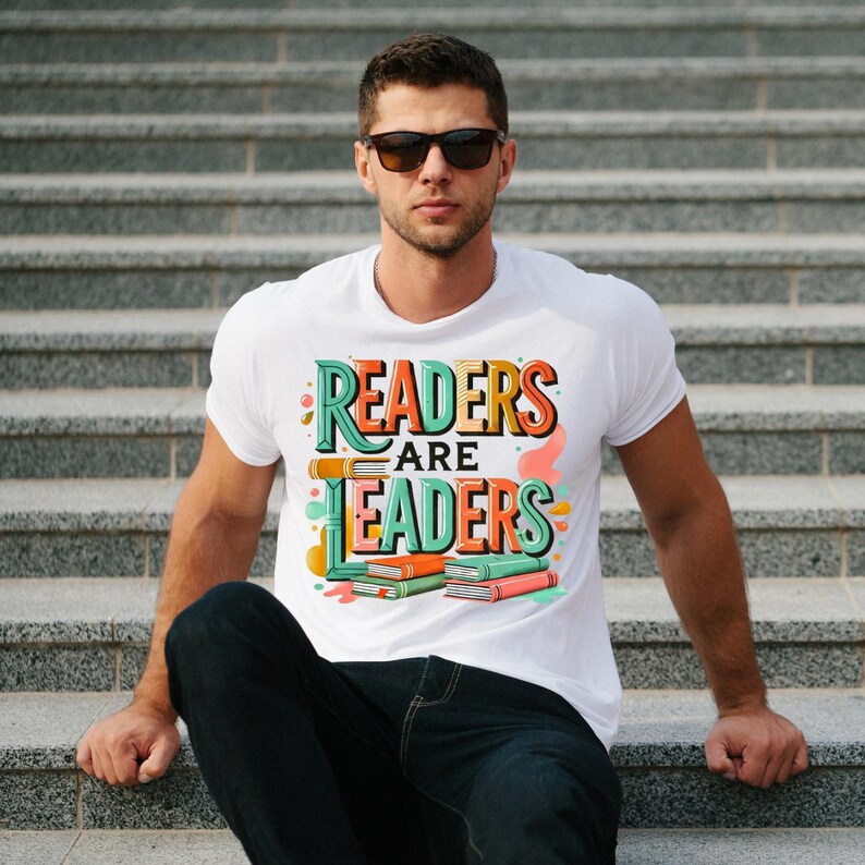 May include: White t-shirt with the text "READERS ARE LEADERS" in a colorful, retro design. The graphic includes stacked books in orange, green, and pink. The person is wearing sunglasses and sitting on gray steps.