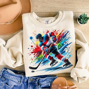 May include: A cream-colored sweatshirt with a colorful graphic of a hockey player skating with a stick and puck. The graphic has splashes of red, blue, green, yellow, and orange paint.