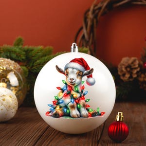 K&ouml;nnte beinhalten: Wei&szlig;er Weihnachtsbaumschmuck mit einer niedlichen Ziege, die einen Weihnachtsmann-Hut tr&auml;gt und in bunte Weihnachtslichter verwickelt ist.