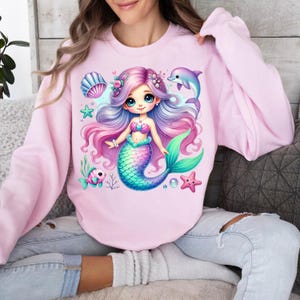 May include: A light pink sweatshirt featuring a colorful mermaid graphic. The mermaid has long, flowing purple and pink hair, a turquoise tail, and is surrounded by sea creatures. A dolphin and starfish are also featured in the design.