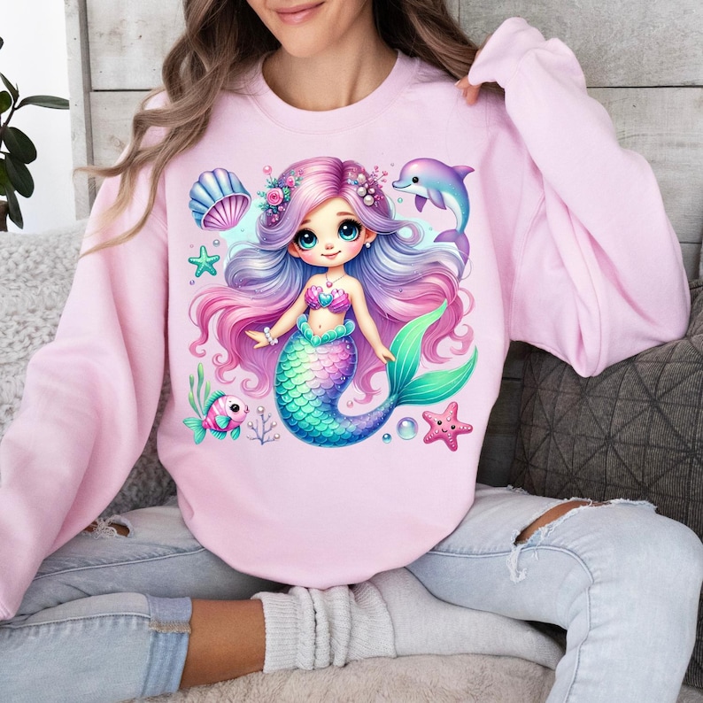 May include: A light pink sweatshirt featuring a colorful mermaid graphic. The mermaid has long, flowing purple and pink hair, a turquoise tail, and is surrounded by sea creatures. A dolphin and starfish are also featured in the design.