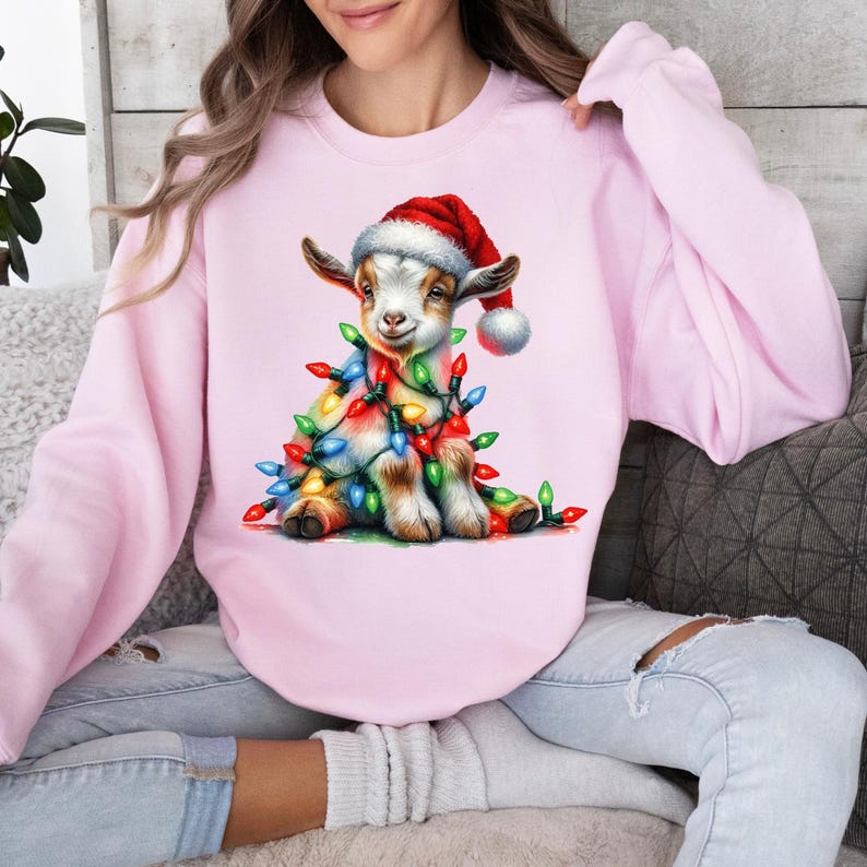 K&ouml;nnte beinhalten: Ein rosa Sweatshirt mit einem niedlichen Cartoon-Ziegenkind, das einen Weihnachtsmann-Hut tr&auml;gt und in Weihnachtslichter gewickelt ist.