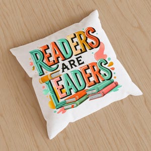 May include: White square pillow with the text "READERS ARE LEADERS" in colorful, stylized lettering. The design includes illustrations of books in shades of orange, green, and red. The pillow is on a light brown wooden surface.