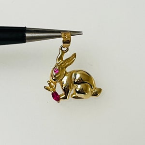 May include: A gold-colored rabbit charm with red gemstone eyes and a red gemstone carrot in its mouth.