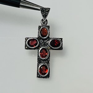 May include: A silver-toned cross pendant with five oval-shaped, deep red gemstones. The cross is detailed with small, dark accents, and is held by a tool. The pendant is likely made of sterling silver and is a religious symbol.