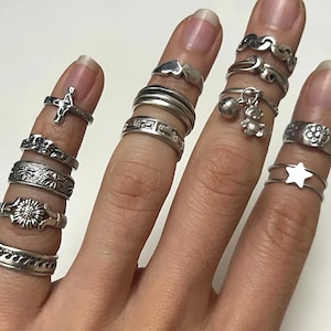May include: A hand wearing a variety of silver rings, including a cross, a flower, a star, and a bear charm.