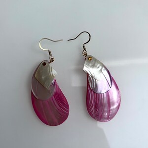 May include: A pair of pink and white shell earrings with a silver hook. The earrings are made of layered shells with a pink and white swirl pattern.