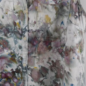 May include: A sleeveless button-down shirt with a floral pattern in shades of white, purple, gray, yellow, and blue. The shirt has two front pockets and a classic collar. The fabric appears to be lightweight.