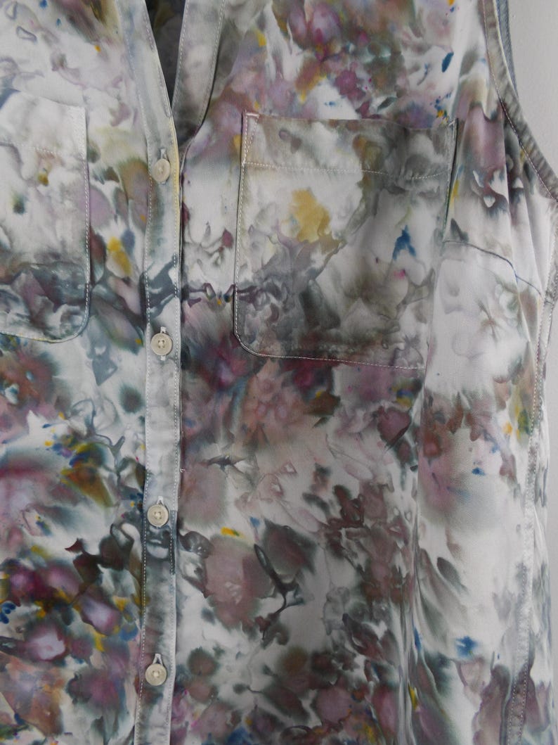 May include: A sleeveless button-down shirt with a floral pattern in shades of white, purple, gray, yellow, and blue. The shirt has two front pockets and a classic collar. The fabric appears to be lightweight.