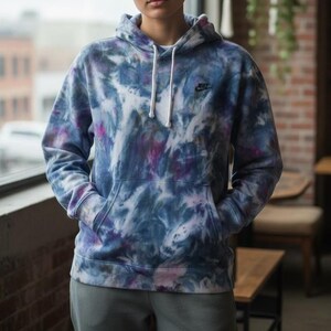 K&ouml;nnte beinhalten: Ein Kapuzenpullover mit Batikmuster in Blau-, Lila- und Wei&szlig;t&ouml;nen. Der Hoodie hat eine Kapuze mit Kordelzug, eine Vordertasche und das Nike-Logo auf der Brust. Das Model tr&auml;gt eine graue Jogginghose.
