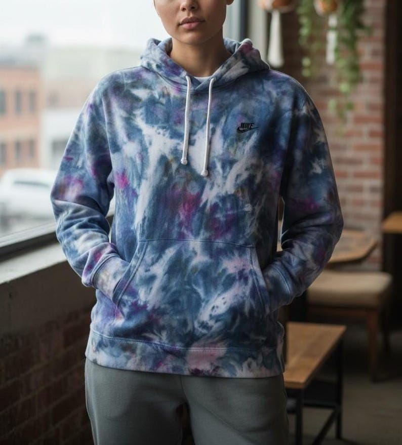 K&ouml;nnte beinhalten: Ein Kapuzenpullover mit Batikmuster in Blau-, Lila- und Wei&szlig;t&ouml;nen. Der Hoodie hat eine Kapuze mit Kordelzug, eine Vordertasche und das Nike-Logo auf der Brust. Das Model tr&auml;gt eine graue Jogginghose.