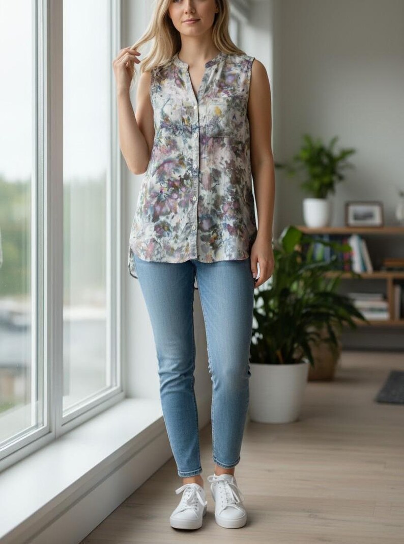 May include: A sleeveless floral print top with a V-neck and button-down front is paired with light blue skinny jeans and white sneakers. The top features a watercolor-like pattern in shades of purple, green, and yellow.