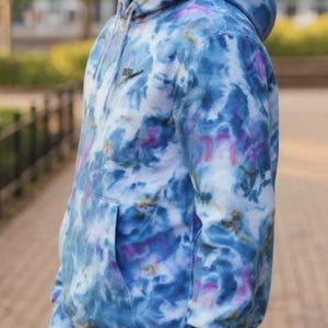 K&ouml;nnte beinhalten: Ein Kapuzenpullover im Batik-Design in Blau-, Lila- und Wei&szlig;t&ouml;nen. Der Hoodie hat eine Vordertasche, eine Kapuze und ein kleines schwarzes Logo auf der Brust. Das Kleidungsst&uuml;ck ist aus einem weichen Material gefertigt.