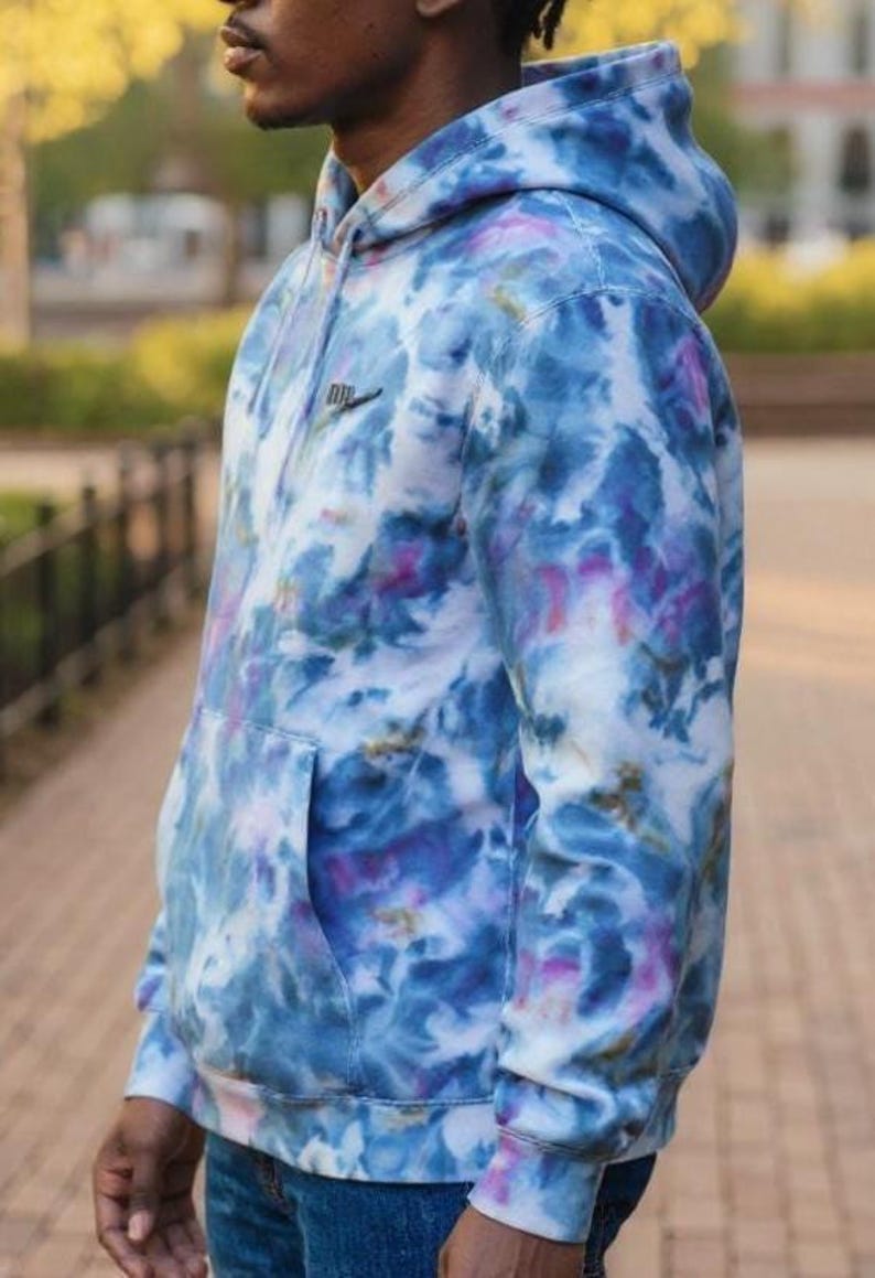 K&ouml;nnte beinhalten: Ein Kapuzenpullover im Batik-Design in Blau-, Lila- und Wei&szlig;t&ouml;nen. Der Hoodie hat eine Vordertasche, eine Kapuze und ein kleines schwarzes Logo auf der Brust. Das Kleidungsst&uuml;ck ist aus einem weichen Material gefertigt.