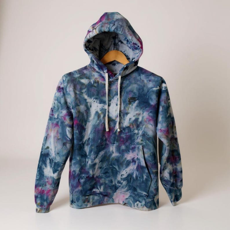 K&ouml;nnte beinhalten: Ein Kapuzenpullover mit Batikmuster in Blau-, Lila- und Rosat&ouml;nen, mit einer Vordertasche und einer Kapuze mit Kordelzug. Das Nike-Logo ist auf der Brust gestickt. Der Hoodie h&auml;ngt an einem Holzkleiderb&uuml;gel.