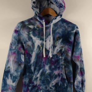 K&ouml;nnte beinhalten: Ein Batik-Hoodie in Blau-, Lila- und Wei&szlig;t&ouml;nen. Der Hoodie hat eine Kapuze mit Kordelzug, eine Vordertasche und das Nike-Logo auf der Brust. Das Kleidungsst&uuml;ck h&auml;ngt an einem Holzkleiderb&uuml;gel.