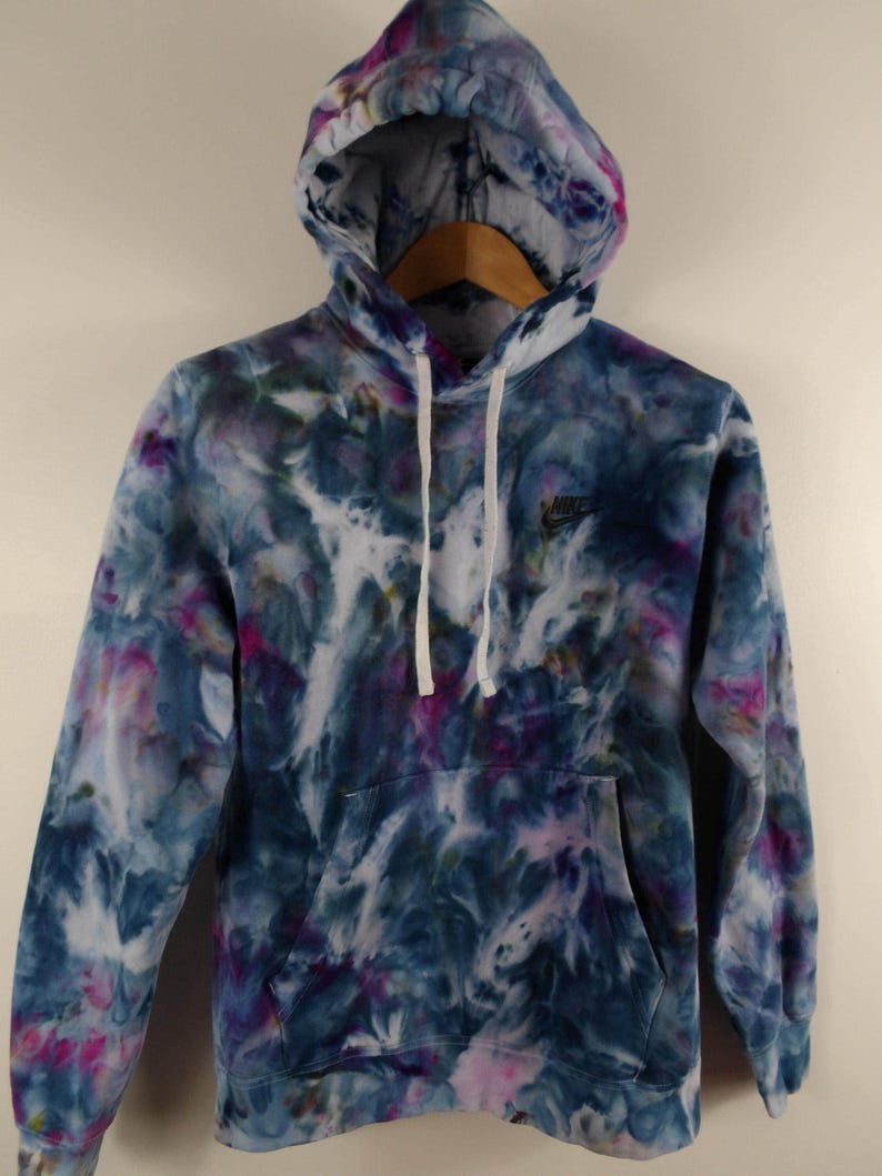 K&ouml;nnte beinhalten: Ein Batik-Hoodie in Blau-, Lila- und Wei&szlig;t&ouml;nen. Der Hoodie hat eine Kapuze mit Kordelzug, eine Vordertasche und das Nike-Logo auf der Brust. Das Kleidungsst&uuml;ck h&auml;ngt an einem Holzkleiderb&uuml;gel.