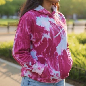 May include: A tie-dye hoodie in shades of pink and light blue. The hoodie features a front pocket and a white Nike logo on the chest. The sleeves have ribbed cuffs, and the hood is lined with light blue fabric.