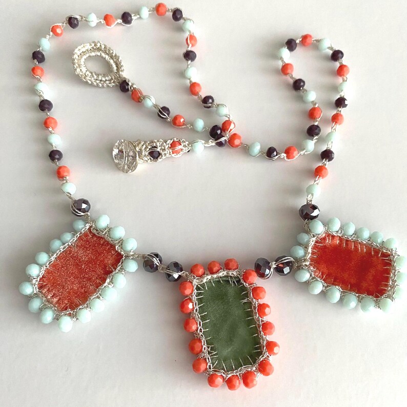 A necklace with three rectangular fabric pieces framed by beads. The fabric pieces are orange, green, and orange. The beads are light blue, orange, and dark purple.