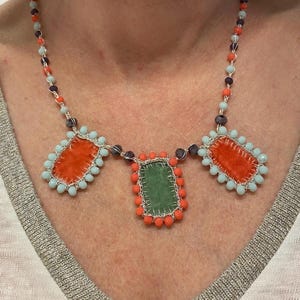 A necklace with three rectangular fabric pieces framed by beads. The fabric pieces are orange, green, and orange. The beads are light blue, orange, and dark purple.