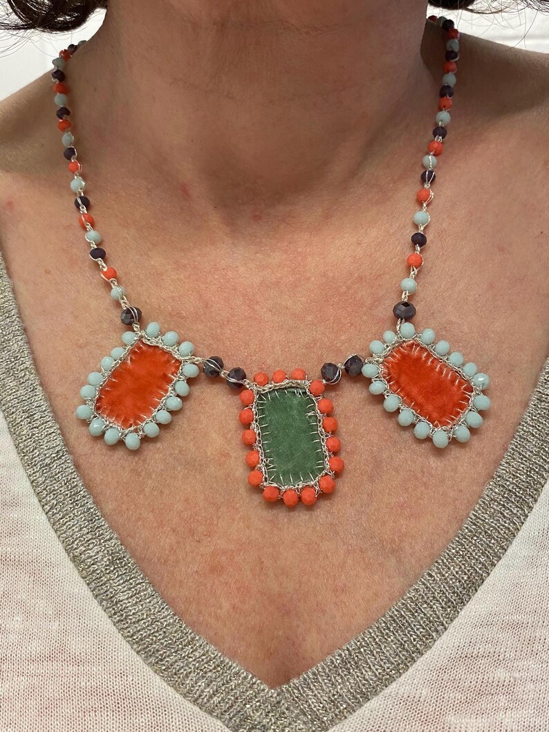 A necklace with three rectangular fabric pieces framed by beads. The fabric pieces are orange, green, and orange. The beads are light blue, orange, and dark purple.