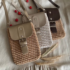 May include: Three crochet crossbody bags in beige, gray, and brown, each with a leather flap and adjustable strap. The bags feature a buckle closure and are displayed on a neutral background.