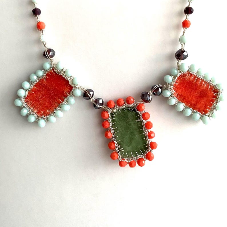 A necklace with three rectangular fabric pieces framed by beads. The fabric pieces are orange, green, and orange. The beads are light blue, orange, and dark purple.