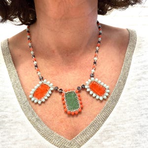 A necklace with three rectangular fabric pieces framed by beads. The fabric pieces are orange, green, and orange. The beads are light blue, orange, and dark purple.