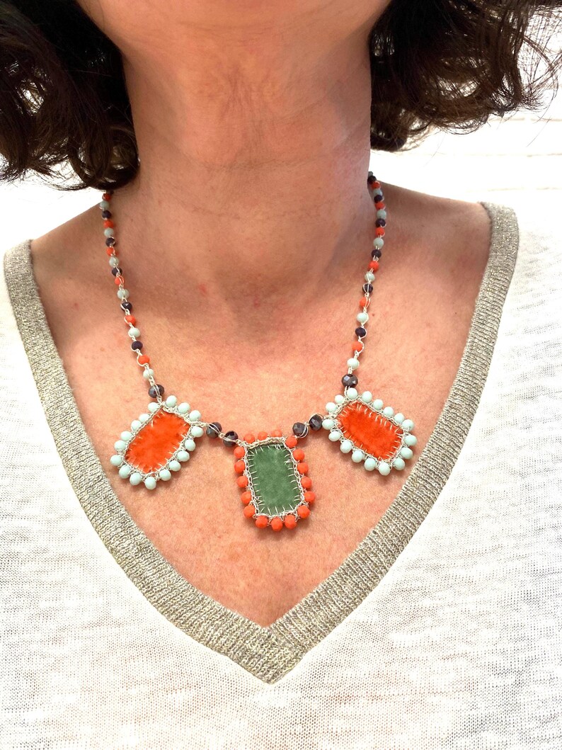 A necklace with three rectangular fabric pieces framed by beads. The fabric pieces are orange, green, and orange. The beads are light blue, orange, and dark purple.