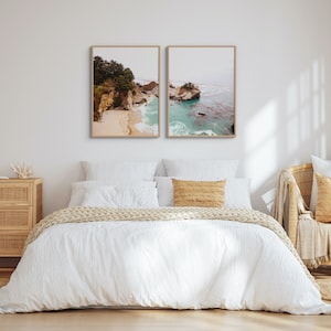 May include: A white bed with a white duvet cover and a tan throw blanket. Two framed photographs of a rocky coastline with turquoise water hang above the bed.