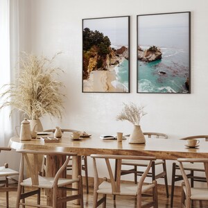 May include: Two framed landscape photographs of a rocky coastline with turquoise water. The photos are hanging on a white wall above a wooden dining table with six chairs.