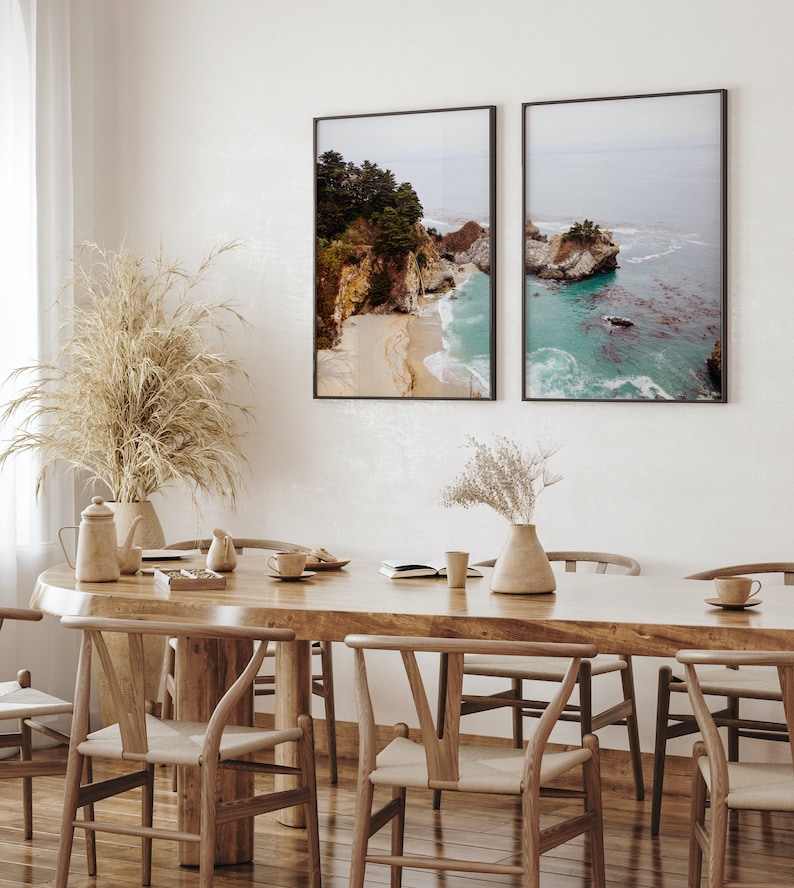 May include: Two framed landscape photographs of a rocky coastline with turquoise water. The photos are hanging on a white wall above a wooden dining table with six chairs.