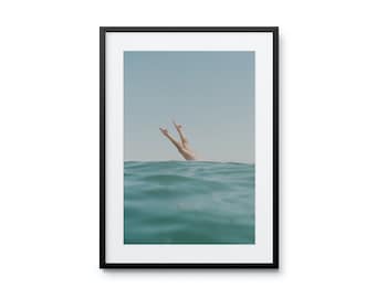 May include: A pair of legs emerge from the water, with the feet pointing upwards towards a blue sky. The water is a vibrant turquoise color.