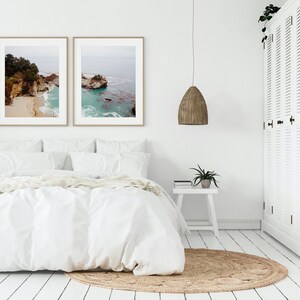 May include: A white bedroom with a bed, a white nightstand, a woven pendant light, and two framed landscape photographs of a rocky coastline.