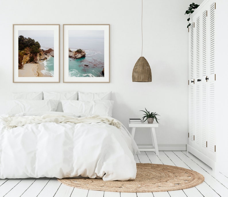 May include: A white bedroom with a bed, a white nightstand, a woven pendant light, and two framed landscape photographs of a rocky coastline.