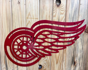 May include: Red metal Detroit Red Wings logo with a wing and a wheel design. The logo is mounted on a wooden surface.